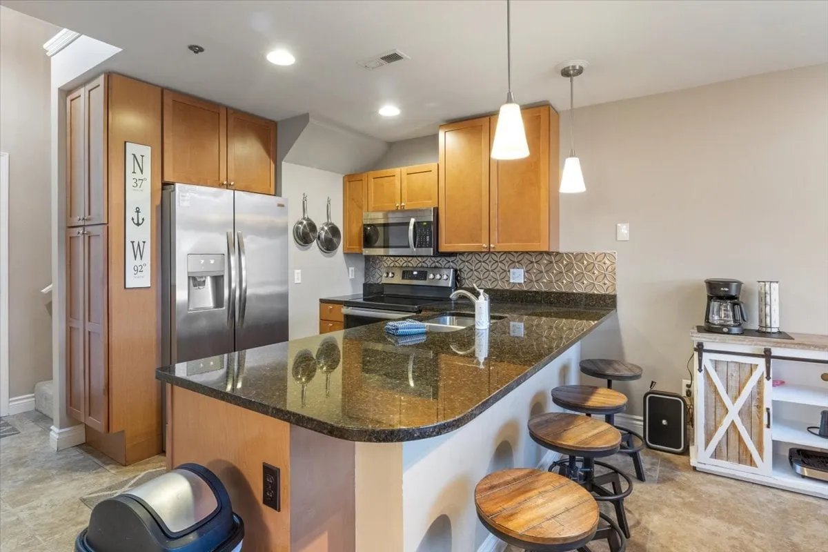 A well-appointed kitchen with granite countertops, a spacious island, stainless steel appliances, wooden cabinets, and pendant lighting. The setup invites family meals and social cooking sessions, reflecting the emphasis on a fully equipped kitchen and gathering space.