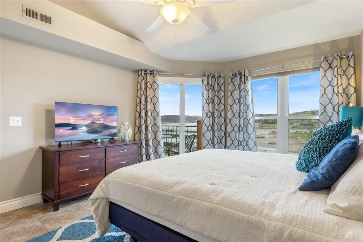 This image features a bedroom with plush bedding, large windows that offer a stunning lake view, a television atop a dresser, and decorative pillows that create a cozy, tranquil atmosphere. The natural light pouring in through the windows highlights the peacefulness and luxury of the master suite.
