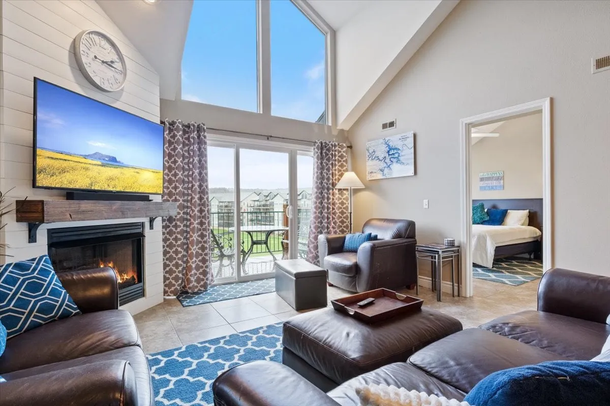A spacious, sunlit living area featuring large windows that frame beautiful lake views. The room is furnished with comfortable seating, a fireplace, a television, and decorative throw pillows, creating a warm and welcoming atmosphere ideal for relaxation and entertainment. The proximity to the balcony hints at easy indoor-outdoor flow, perfect for soaking in sunsets or enjoying a cozy movie night.