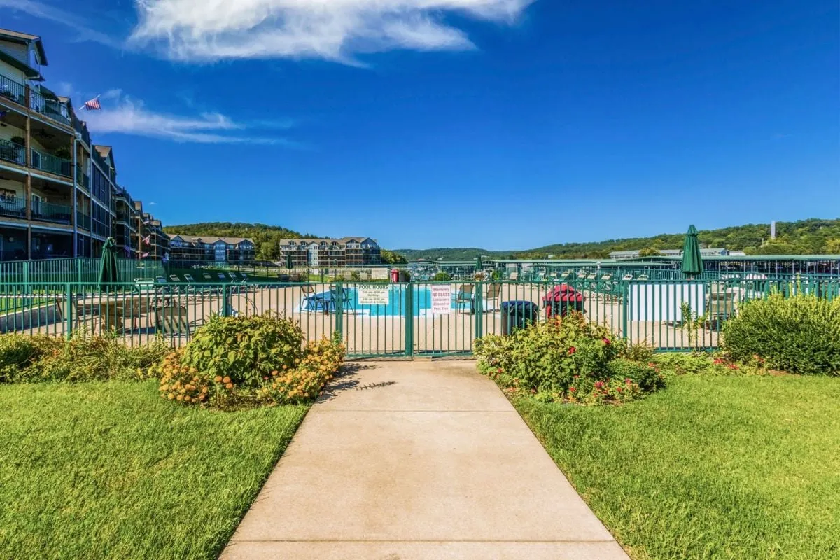 An exterior scene showcasing a swimming pool area with lounge chairs, a gated entrance, manicured lawns, and a waterfront view. The image reflects the community's resort-style amenities and highlights the proximity to the water, crucial for fishing and boating activities.