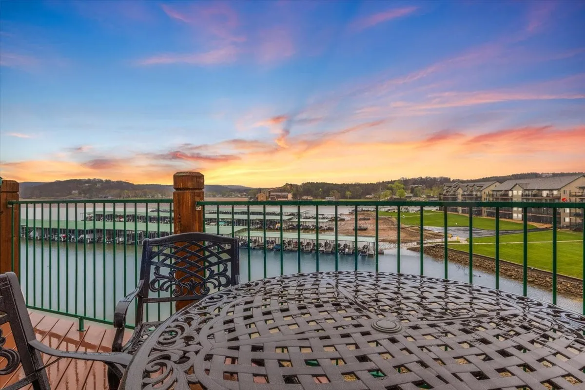 The image showcases a spacious outdoor dining area with a table and chairs overlooking a tranquil stretch of water. The sky is painted with the warm hues of sunset, creating a peaceful and inviting atmosphere ideal for gathering with family and friends. The waterfront view highlights the condo's proximity to Lake of the Ozarks and sets a serene scene for relaxation, dining, and socializing.