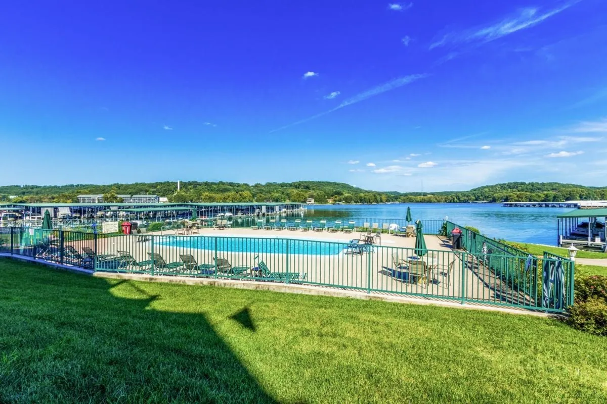 A sparkling swimming pool surrounded by comfortable lounge chairs, with a scenic lake view and well-kept green grass enclosed by a safe fence. The relaxed mood hints at both sunbathing and social gatherings by the water.