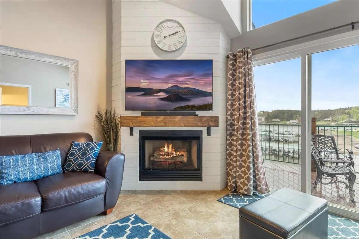 A cozy living area with a fireplace, a TV displaying a scenic landscape, a comfortable sofa, curtains, and a balcony view that hints at the natural beauty outside. The space offers warmth and comfort, with a glimpse of the outdoors visible through the windows and balcony.